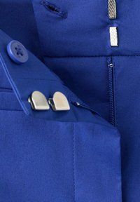 Gros plan d'un vêtement bleu avec bouton, fermeture éclair, boutonnière et deux fermetures à crochets et barres en métal argenté sur une couche de tissu.