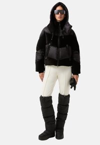 Sort polstrede jakke med hætte, lynlåsede lommer og blank overflade; parret med hvide tætsiddende bukser og sorte isolerede støvler.