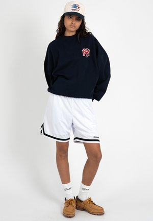 Mladý člověk nosící tmavě modrou mikinu s logem NY, bílé basketbalové šortky, bílé ponožky, hnědé boty a béžovou baseballovou čepici s designem.