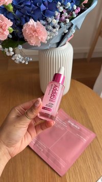 Rosa Glasserumflasche mit Pipette, verziert mit silbernen Akzenten. Der Hintergrund zeigt ein blaues und pinkes Blumenarrangement sowie eine helle Holzoberfläche.