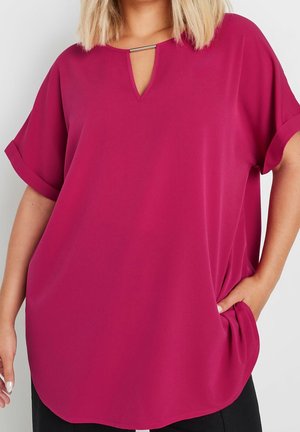 Femme portant un haut ample magenta avec des manches courtes retroussées et un petit détail en barre métallique au col en V, main dans la poche côté droit.