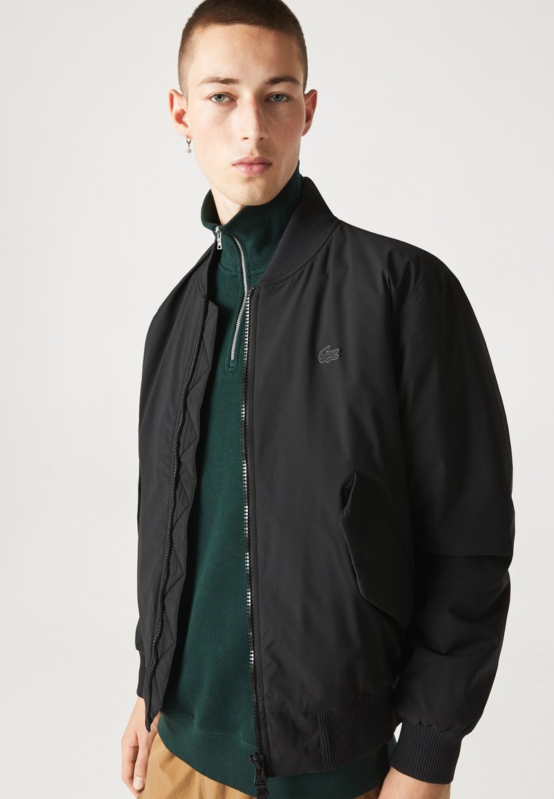 Blouson noir avec fermeture éclair à l'avant, poignets côtelés et logo Lacoste. Porté par-dessus un haut vert foncé. Tissu lisse avec une finition mate.