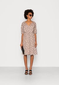 Veste rosa clara com estampa floral, decote em V, mangas curtas em balão, cintura com cinto e saia evasé. Usada com acessórios pretos e óculos de sol.