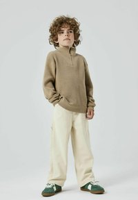 Beżowy sweter z wysokim kołnierzem, zestawiony z luźnymi kremowymi spodniami cargo. Całość dopełniają zielono-białe sneakersy.