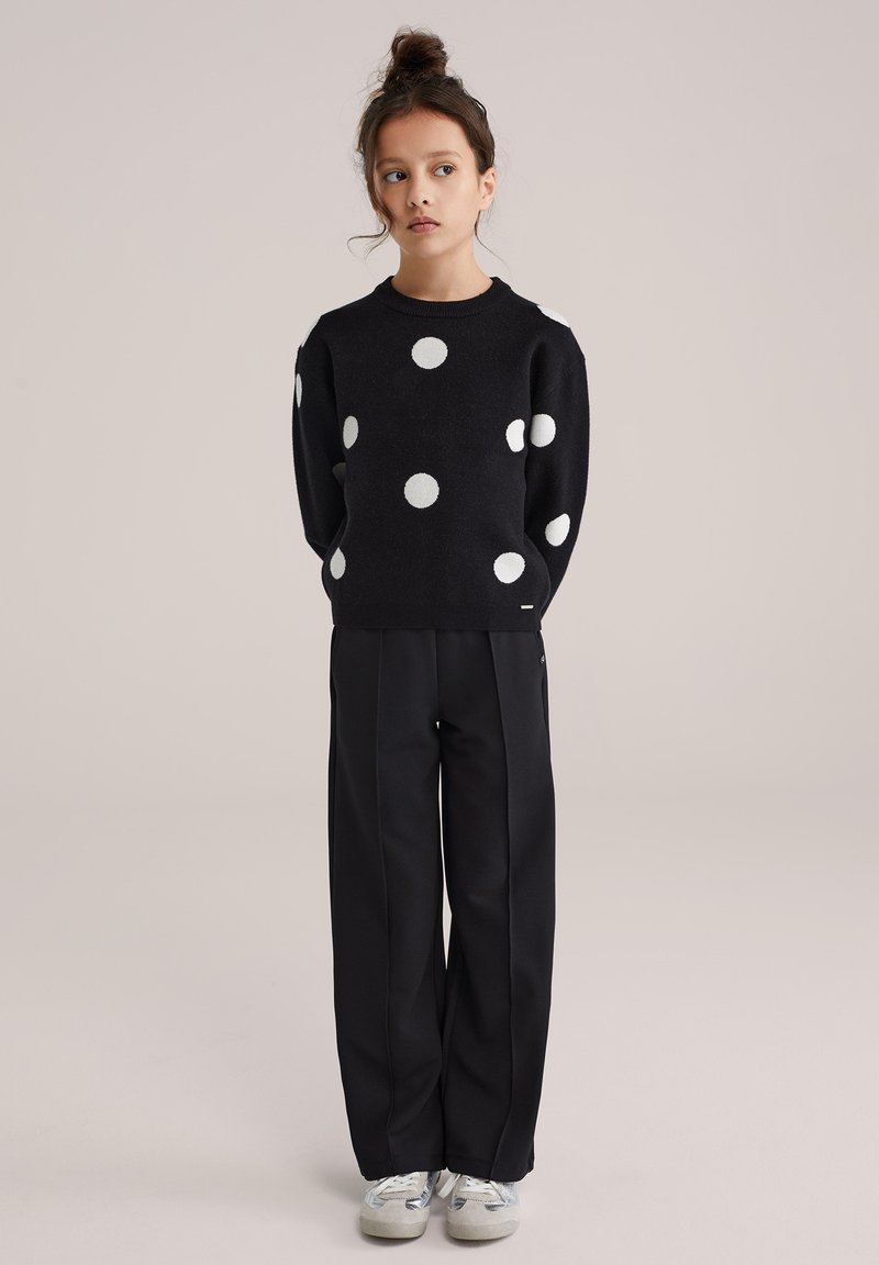 Pull noir à gros pois blancs, col rond, associé à un pantalon large noir et à des baskets grises. Texture tricotée.