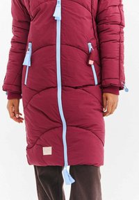 Burgundská dlouhá péřová bunda s modrými zipy, dvěma bočními kapsami a žebrovanými manžetami. Obsahuje prošívaný vzor a malou textilní štítek.