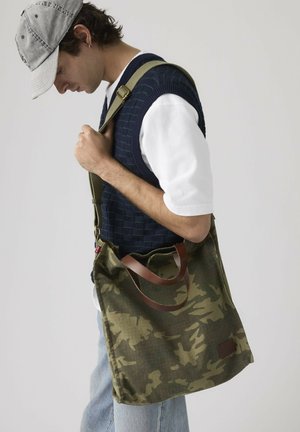 Jonge man met een grijze pet, navy blauwe gebreide spenser over een wit shirt, met een groene camouflage schoudertas met bruine leren handvatten.