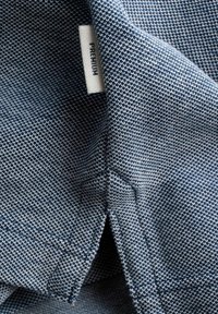 Gros plan sur un tissu texturé bleu avec une étiquette blanche "PREMIUM" cousue près d'un ourlet piqué et un détail d'ouverture.