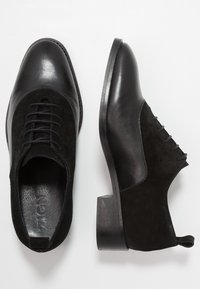 Chaussures en cuir noir et en daim avec un bout rond, un laçage, et un petit talon empilé. L'empeigne combine des matériaux lisses et texturés.