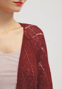 Röd stickad cardigan med öppen design, prydd med ett spetsmönster, långärmad och med en mjuk textur, bärs över en ljusrosa topp.