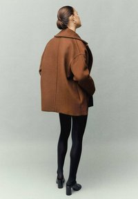Abrigo de lana marrón oversized con un collar ancho, diseño minimalista y detalle en la costura trasera, combinado con medias negras y zapatos de tacón cuadrado.
