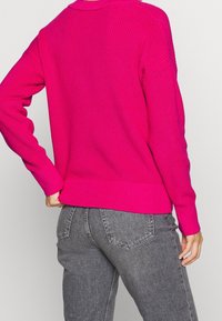 Fuchsia gestrickter Pullover mit Rippstrick-Textur und langen Ärmeln. Hat einen runden Halsausschnitt und eine lässige Passform. Kombiniert mit grauen Jeans.