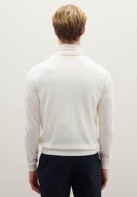 Hellcremiger Pullover mit hohem Kragen, gerippten Bündchen und Saum; weiche Textur; Modell schaut weg; kombiniert mit dunkler Hose.