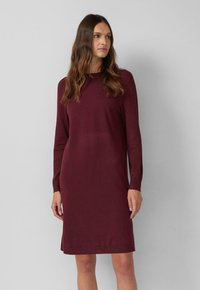 Frau steht, trägt ein knielanges, langärmliges, kastanienbraunes Strickkleid und schaut zur Seite vor einem schlichten hellgrauen Hintergrund.