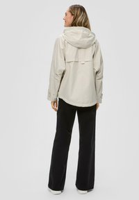 Lichtbeige jas met capuchon, getailleerde taille en een verlengde achterkant. Bevat een rugventilatie en drukknoopmanchetten. Gekocht bij een zwarte broek en sneakers.