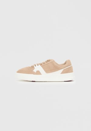 Suede sneakers in beige with white accents. Rounded toe, lace-up closure, and a thick white sole with a hint of red at the bottom.