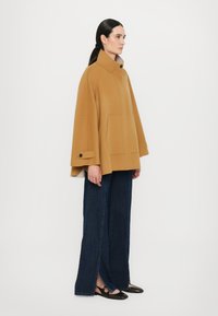 Cappa in misto lana color cammello con un colletto alto, tasche laterali e maniche con bottoni, abbinata a pantaloni a gamba larga in denim scuro.