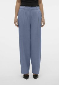 Wijde broek in lichtblauwe stof, met een textuurafwerking, voorplooien, riemlussen en zwarte puntige schoenen.