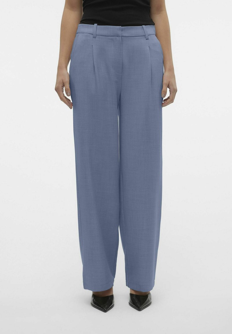 Wijde broek in lichtblauwe stof, met een textuurafwerking, voorplooien, riemlussen en zwarte puntige schoenen.