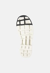 Semelle de chaussure blanche avec un motif texturé ; présente un recouvrement en caoutchouc noir à l'avant avec des découpes en forme de grille et une indication de taille 36.