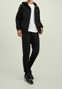 Felpa nera con cappuccio e lacci, tasche frontali, abbinata a pantaloni della tuta neri e sneakers nere con accenti multicolori. T-shirt bianca sotto.