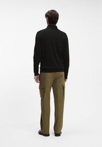 Άνδρας στέκεται με την πλάτη προς τα εμάς, φοράει μαύρο πουλόβερ με μακριά μανίκια και χακί cargo παντελόνι με τσέπες με καπάκι, μπροστά σε απλό λευκό φόντο.