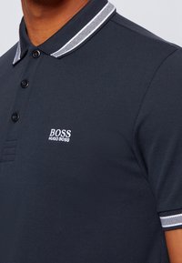 Polo shirt bleu foncé en tissu texturé, avec un col à rayures contrastées, une patte de boutonnage à trois boutons et un logo brodé blanc.