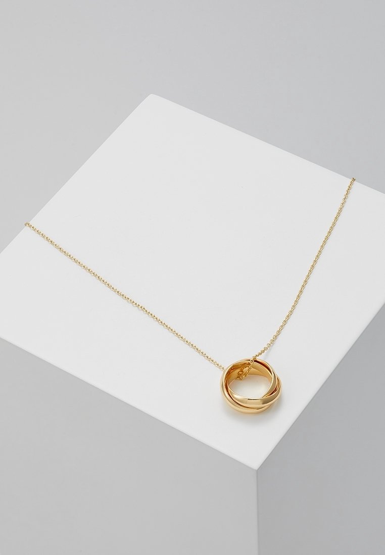 Collier chaîne en or avec trois anneaux en or polis et entrelacés exposés sur un bloc géométrique blanc sur un fond gris.