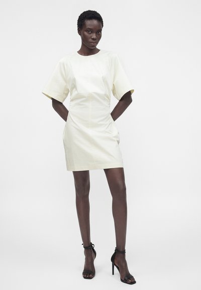 Γυναίκα που φοράει ένα κοντομάνικο, εκρού φόρεμα με στενή μέση, ποζάροντας με τα χέρια πίσω από την πλάτη και φορώντας μαύρα ψηλοτάκουνα σανδάλια.