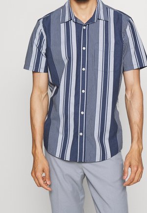 La chemise à manches courtes présente des rayures verticales bleu marine et bleu clair, un devant à boutons et une petite poche sur la poitrine en tissu doux.