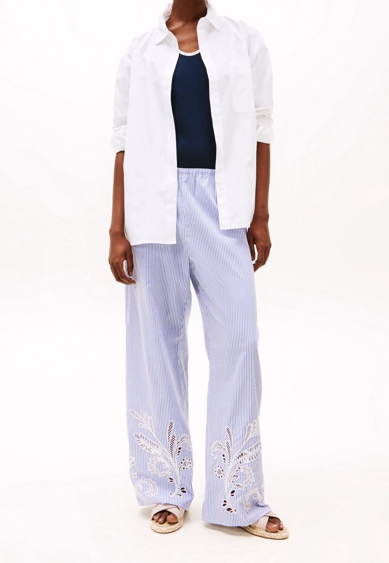 Chemise blanche à boutons, débardeur bleu marine, pantalon large à rayures bleu avec broderie en dentelle à l'ourlet, et espadrilles de couleur naturelle.