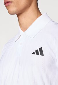Biała koszulka polo z fakturowaną tkaniną, standardowym kołnierzykiem i czarnym logo z trzema ukośnymi paskami na lewej piersi.
