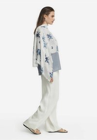 Poncho floral léger en blanc et bleu, superposé sur un haut gris, associé à un pantalon blanc et des sandales accentuées de silver.
