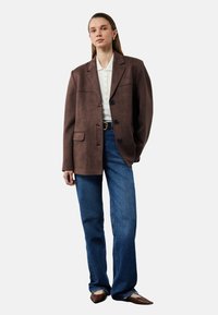 Brauner Wildleder-Blazer mit einer Brusttasche und drei Knöpfen, kombiniert mit hellblauen, weit geschnittenen Jeans und spitzen braunen Schuhen.