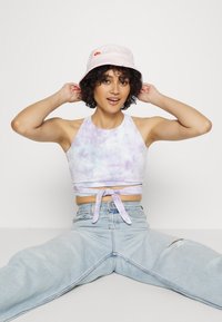 Top cropped roxo em tie-dye com detalhe de nó na frente, combinado com jeans mom claros e desfiados. Inclui um chapéu de balde rosa claro com logo.