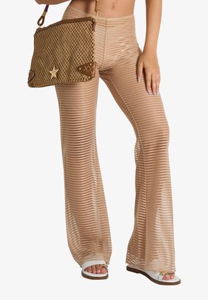 Donna che indossa pantaloni a campana beige trasparenti a righe e sandali bianchi, tenendo una borsa intrecciata marrone con dettaglio a stella.