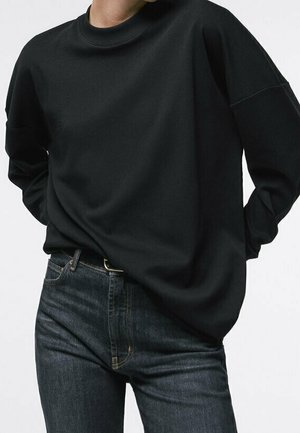 Person trägt ein lockeres schwarzes Langarmshirt, das in hoch taillierte dunkelblaue Jeans gesteckt ist, mit den Händen hinter dem Rücken.