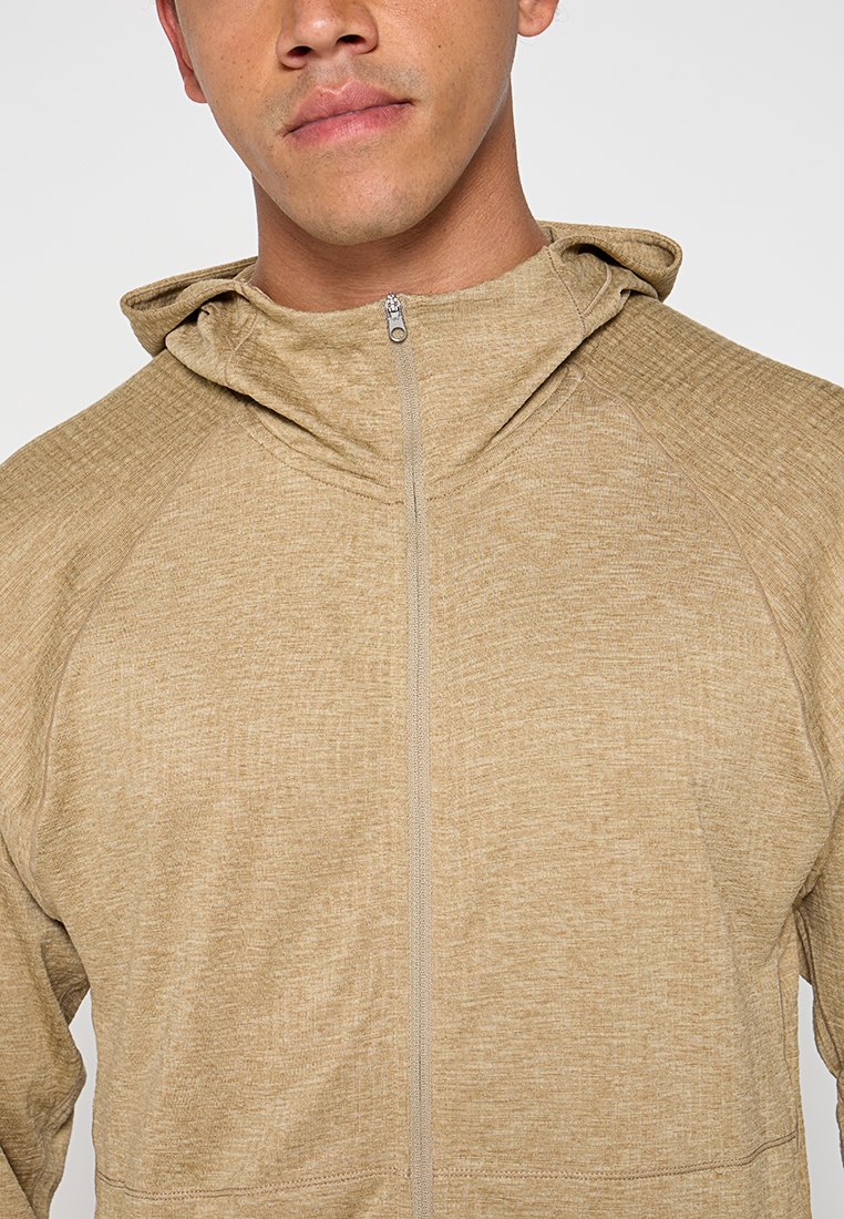 Uomo che indossa una felpa con zip di colore marrone chiaro con la zip completamente chiusa su uno sfondo neutro.