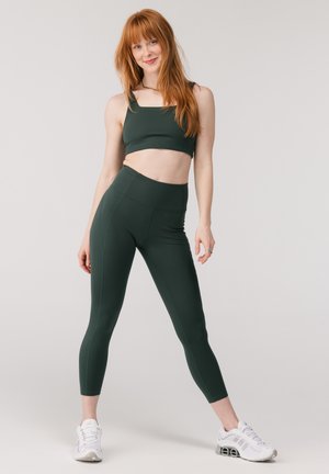 Joven mujer con pelo rojo largo que lleva un sujetador deportivo verde oscuro y mallas, sonriendo y con las piernas separadas contra un fondo liso.