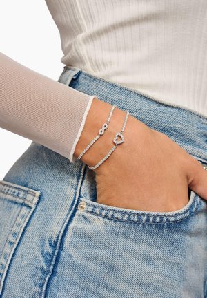 Bracelets en argent ornés de pierres claires, incluant un design en forme de cœur et d'infini, portés sur une main près d'une poche en denim avec un haut de couleur claire.