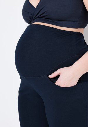 Schwangere Frau in marineblauer Umstandshose mit der Hand in der Tasche, trägt ein passendes marineblaues Crop-Top vor hellem Hintergrund.