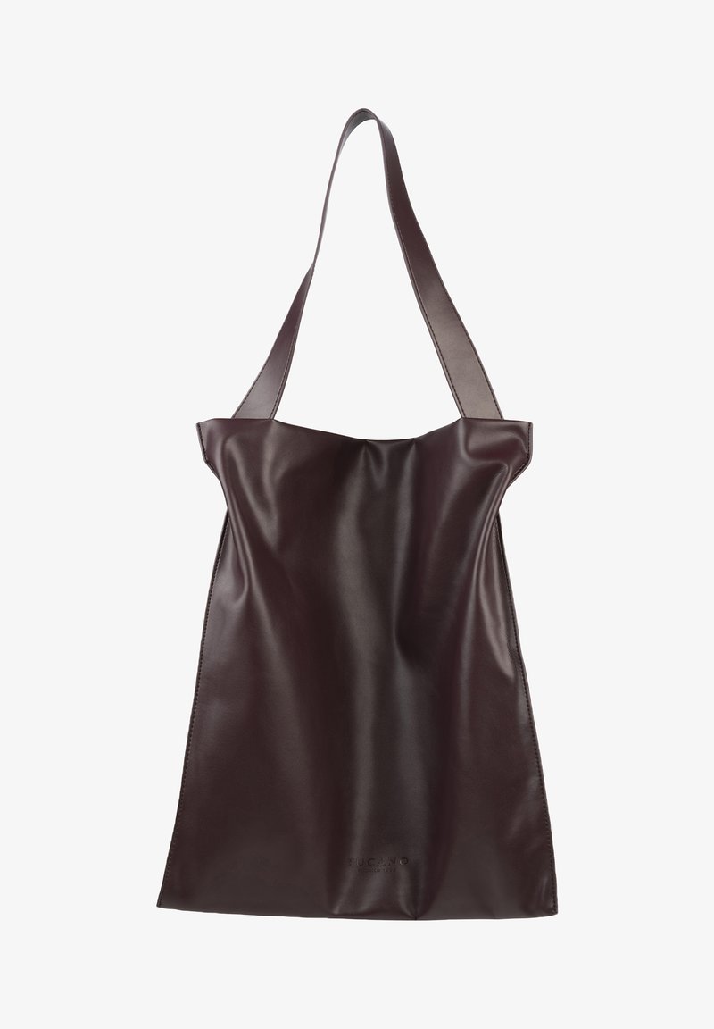 Leren totebag in donkerbruin. Gladde textuur, oversized vorm met brede schouderbanden, en een subtiel logo dat bij de onderkant is gedrukt.