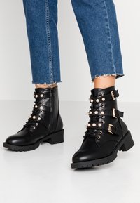 Černé kožené kotníkové boty s robustním podpatkem, zdobené detaily přezek, perlovými akcenty a šněrováním vpředu. Texturovaná gumová podrážka.