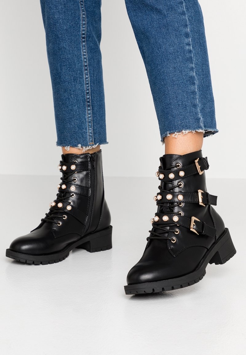 Černé kožené kotníkové boty s robustním podpatkem, zdobené detaily přezek, perlovými akcenty a šněrováním vpředu. Texturovaná gumová podrážka.