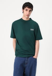 Grønn bomull T-skjorte med rund hals, kortermet, og liten hvit trykt tekst "the new originals" på venstre bryst. Kombinert med blå jeans.