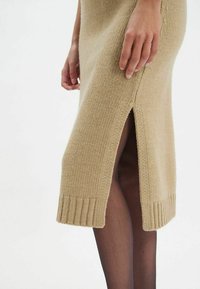 Robe en tricot beige avec une fente latérale, un ourlet côtelé et un tissu texturé. Gros plan sur l'ourlet et une partie du bras visible sur un fond uni.