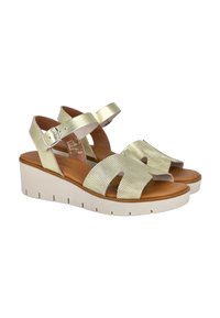 Sandalias de cuña con una correa metálica de color dorado claro y un diseño perforado, que presentan una plantilla de cuero marrón y una suela blanca gruesa.