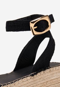 Espadrille en textile noir avec détails en métal doré. Semelle en jute tressé avec une semelle intérieure noir lisse. Présente un design à lanières.