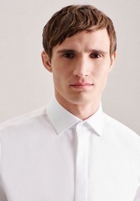 Camicia bianca con colletto a punte e bottoni sul davanti, caratterizzata da una texture liscia e una vestibilità classica. Design minimalista senza motivi o dettagli.