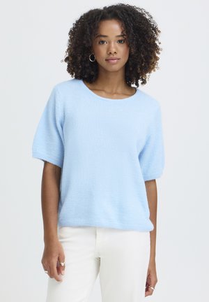 Jeune femme aux cheveux bouclés portant un pull à manches courtes bleu clair et un pantalon blanc, debout devant un fond uni.